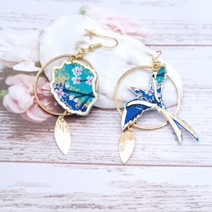 Può includere: Un paio di orecchini a cerchio dorati con un design floreale blu e verde. Un orecchino presenta un design a forma di uccello, mentre l'altro presenta un design floreale.
