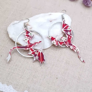 Op de afbeelding: Een paar zilveren creolen met rode drakenbedels. De draken zijn gemaakt van gepatenteerd stof met witte bloemen en hebben zilveren vleugels.