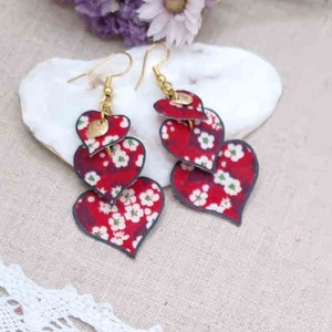 May include: A pair of dangle earrings with three layered red hearts with a white floral pattern. The hearts are outlined in black and have gold accents.