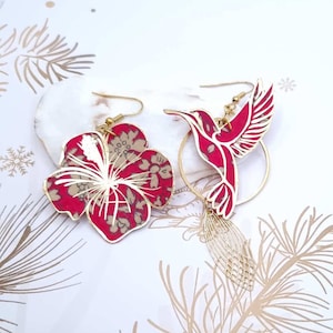 May include: A pair of gold earrings featuring a red hibiscus flower and a red hummingbird design. The earrings are crafted from metal and have a delicate, feminine aesthetic.