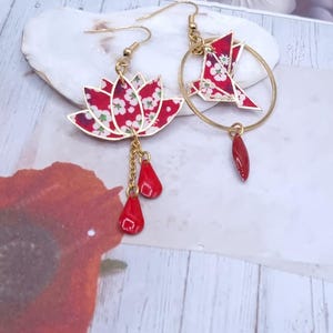 May include: Gold-tone earrings featuring a red and white floral lotus flower and a red origami bird within a gold hoop. The lotus has red teardrop charms, and the bird has a red teardrop charm. The earrings are on a white and beige background.