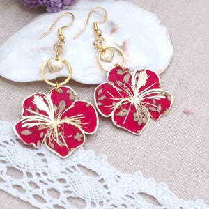 Peut inclure: Une paire de boucles d'oreilles en or avec des breloques de fleurs d'hibiscus rouges. Les fleurs ont un centre blanc et sont détaillées avec un motif floral doré.