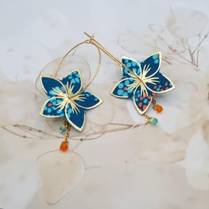 Könnte beinhalten: Goldene Creolen-Ohrringe mit blauen Blumenanhängern. Die blütenförmigen Anhänger haben einen goldenen Umriss und sind mit einem blau-orangen Muster verziert. Kleine orangefarbene, blaue und grüne Perlen hängen von der Unterseite jedes Ohrrings.