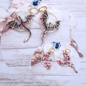 May include: Dragon-shaped earrings with gold-colored hoops. The dragon wings are red and black with gold accents. The earrings are displayed on a white surface with floral and wooden background.