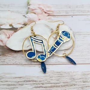 Peut inclure: Une paire de boucles d'oreilles dorées. L'une présente une note de musique bleue, l'autre un motif de microphone bleu. Les deux boucles d'oreilles ont un petit charme en forme de goutte d'eau bleu foncé.