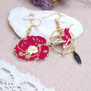 May include: A pair of gold earrings with a red floral design and a hummingbird design. The floral earring has a red and white floral pattern. The hummingbird earring has a gold circle with a black bead hanging from it.