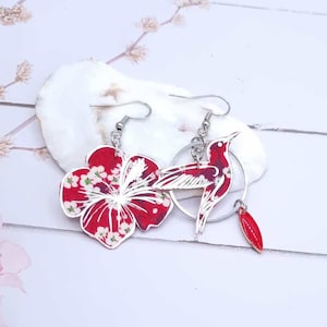 Puede incluir: Un par de pendientes colgantes con una flor de hibisco roja y un colibrí con un acento rojo. Los pendientes están hechos de metal y tienen un acabado plateado.