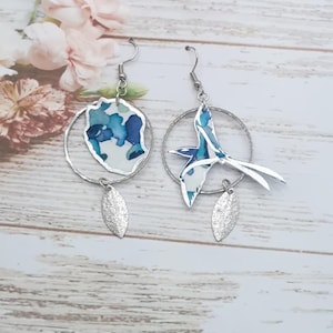 May include: A pair of silver earrings. One earring has a circular design with blue and white watercolour patterns. The other has a bird-shaped design with similar blue and white patterns. Both earrings have a silver leaf charm.