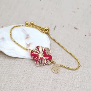 May include: A gold chain bracelet with a red and beige hibiscus flower charm and a gold monstera leaf charm.