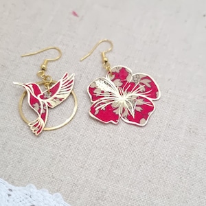 May include: A pair of gold earrings featuring a hummingbird and a red hibiscus flower design. The hummingbird is in flight and the hibiscus flower has a white centre.