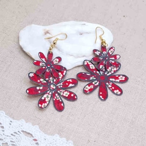 Può includere: Un paio di orecchini pendenti a forma di fiore rosso con ganci dorati. Gli orecchini sono realizzati con fiori di carta sovrapposti con un motivo floreale bianco.