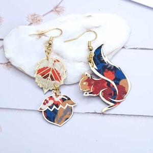 Puede incluir: Un par de pendientes colgantes de oro con una ardilla roja y azul y una bellota marrón con un dije de hoja de oro. Los pendientes están hechos de tela y tienen un patrón floral.