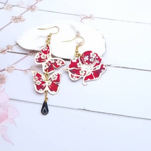 May include: A pair of dangle earrings featuring a red and white floral pattern. One earring has a single flower design, while the other has two butterfly designs with a black teardrop charm.