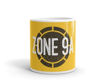 May include: A yellow ceramic mug with a black and white graphic of a circle with the text "ZONE 9A" inside.