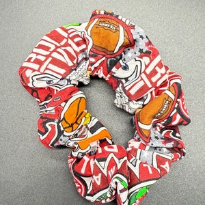 Pode incluir: Uma scrunchie de tecido com um padrão repetido de bolas de futebol americano e logotipos de equipas em vermelho, branco e preto. A scrunchie é circular e parece ser feita de um material macio e flexível. O fundo é cinzento liso.