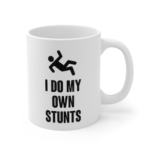 Puede incluir: Taza de cerámica blanca con un gráfico negro de una persona cayendo y el texto "I DO MY OWN STUNTS" en letras negras. La taza tiene un asa curvada.