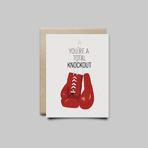 May include: Greeting card with a beige envelope. The card features red boxing gloves hanging from a string, with the text "YOU'RE A TOTAL KNOCKOUT" in black. The background is a light beige.