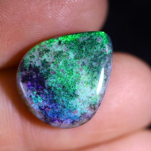 Könnte beinhalten: Ein polierter Opal-Edelstein mit einer lebendigen Farbgebung. Der Stein weist eine Mischung aus grünen, blauen und violetten Farbtönen mit einem gesprenkelten Muster auf seiner Oberfläche auf. Der Opal wird in einer Hand gehalten und zeigt seine einzigartige Form und schillernden Eigenschaften.