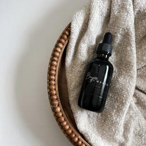 May include: A black glass bottle with a dropper, labeled "Boujee Pumpkin," rests on a beige textured cloth inside a wooden beaded tray. The bottle has a black cap and a white label. The tray is oval-shaped and has a light brown color.