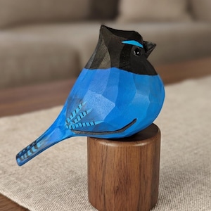 Puede incluir: Escultura de pájaro de madera tallada a mano. El pájaro tiene un cuerpo azul, cabeza negra y una pequeña percha de madera. La escultura está detallada con patrones de plumas talladas y un pequeño acento azul sobre el pico. El pájaro está posado sobre una base cilíndrica de madera.