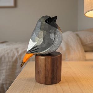以下が含まれることがあります： 手彫りの木製鳥の彫刻。グレーと黒の体、白い翼のアクセント、オレンジ色の尾があります。鳥は円筒形の木製の台座に止まっています。装飾品です。