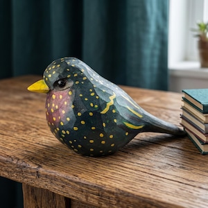 Side profile of a hand-carved wooden European Starling figurine showcasing its intricate, hand-painted yellow speckles over glossy dark iridescent green and purple plumage. The realistic basswood bird sculpture features a bright yellow beak and sits perched on a rustic wood surface next to a decorative stack of books. Perfect for nature-inspired home decor, office desk styling, or a unique handmade gift for bird watchers.
