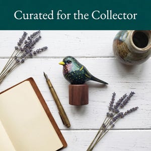 Side profile view of a hand-carved wooden European Starling figurine showcasing its intricate, hand-painted yellow speckles over glossy dark iridescent green and purple plumage. The realistic basswood bird sculpture is perched on a round black walnut base, styled on a white wood surface next to a leather sketchbook, a classic fountain pen, and dried lavender sprigs. Perfect for nature-inspired home decor, office desk styling, or a unique gift for bird watchers.