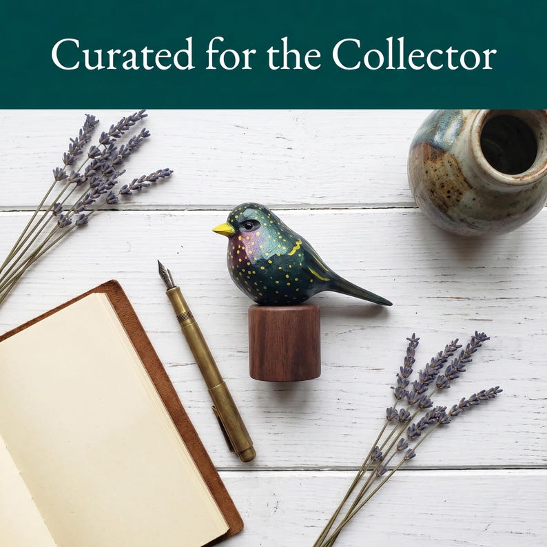 Side profile view of a hand-carved wooden European Starling figurine showcasing its intricate, hand-painted yellow speckles over glossy dark iridescent green and purple plumage. The realistic basswood bird sculpture is perched on a round black walnut base, styled on a white wood surface next to a leather sketchbook, a classic fountain pen, and dried lavender sprigs. Perfect for nature-inspired home decor, office desk styling, or a unique gift for bird watchers.