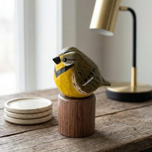 Hand-carved wooden Western Meadowlark figurine with a faceted whittled texture and hand-painted yellow and black plumage. The bird sculpture is displayed on a wooden table next to a gold lamp and stone coasters, perfect for rustic home decor, a nature-themed shelf sitter, or a unique gift for bird watchers.