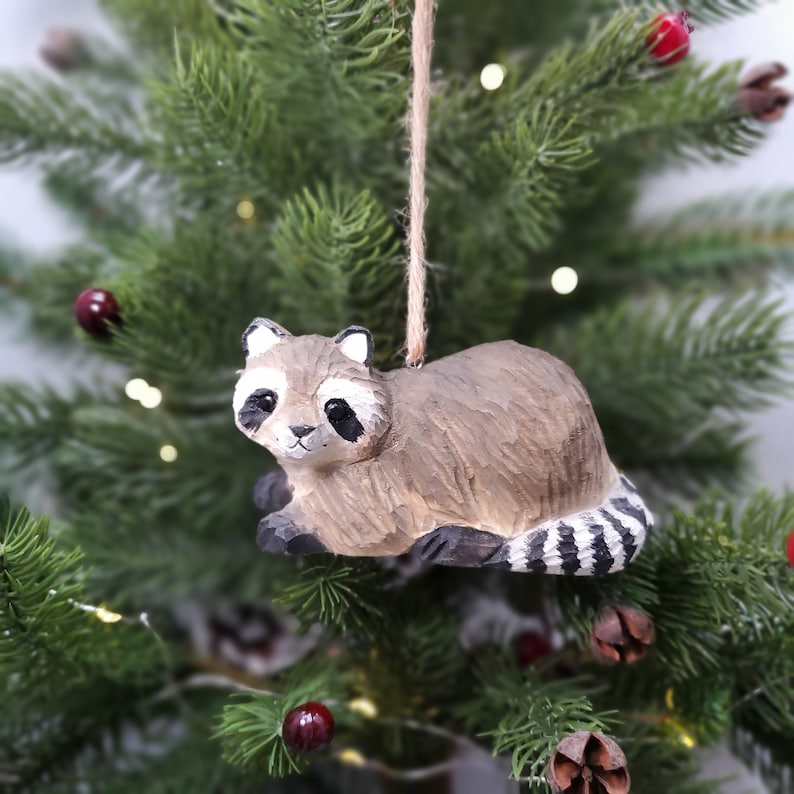 K&ouml;nnte beinhalten: Ein h&ouml;lzerner Waschb&auml;r-Anh&auml;nger mit schwarz-wei&szlig;en Streifen am Schwanz, der an einer braunen Schnur an einem gr&uuml;nen Weihnachtsbaum h&auml;ngt.