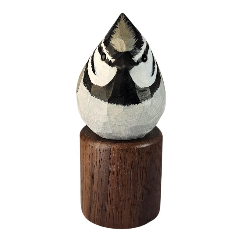 May include: A hand-carved wooden bird figurine. The bird is painted in shades of white, gray, and black, with a detailed face. It is mounted on a dark brown wooden base. The figurine is a decorative item.
