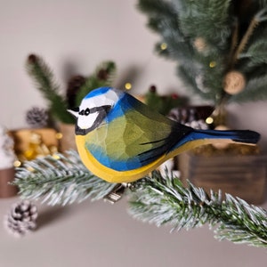 Pode incluir: Figurinha de pássaro de madeira esculpida à mão, um chapim-azul, pousada num ramo de pinheiro nevado. O pássaro apresenta cabeça azul, peito amarelo e dorso verde. A figurinha é presa a um clipe, adequada como enfeite.