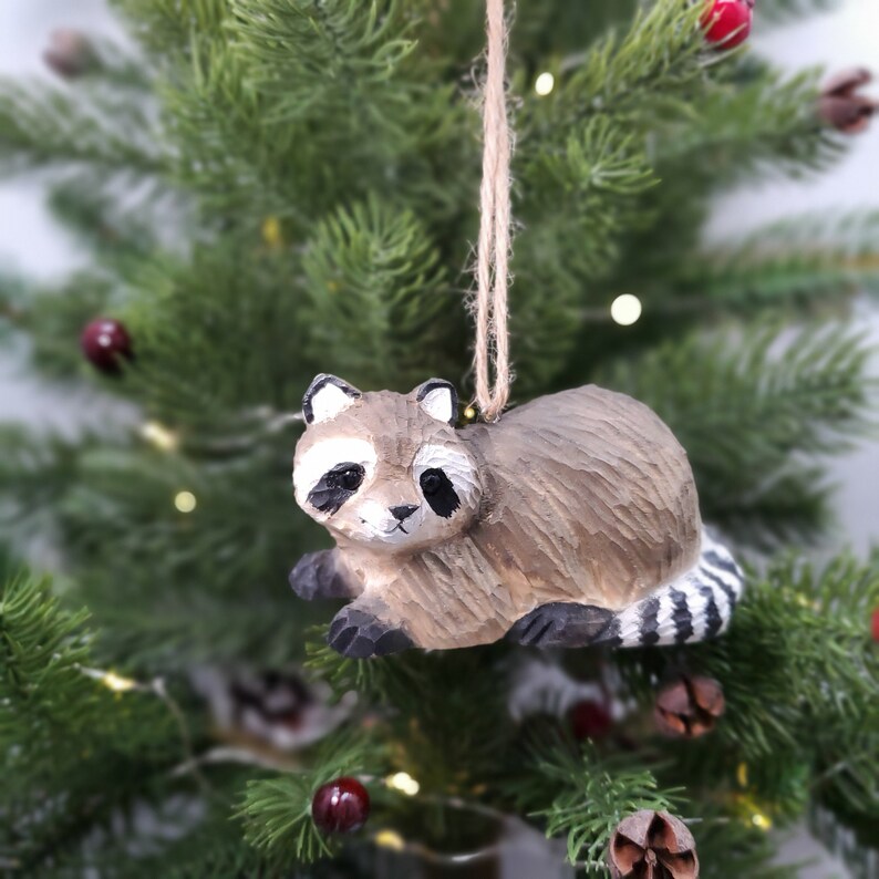 K&ouml;nnte beinhalten: Ein h&ouml;lzerner Waschb&auml;r-Anh&auml;nger mit schwarz-wei&szlig;en Streifen am Schwanz. Der Anh&auml;nger h&auml;ngt an einer braunen Schnur an einem gr&uuml;nen Weihnachtsbaum.