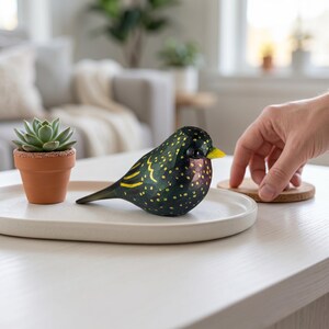 Side profile view of a hand-carved wooden European Starling figurine showcasing its intricate, hand-painted yellow speckles over glossy dark iridescent green and purple plumage. The realistic basswood bird sculpture features a bright yellow beak and is staged on a modern white ceramic plate next to a small terracotta potted succulent under warm light. Perfect for nature-inspired home decor, office desk styling, or a unique handmade gift for bird watchers.