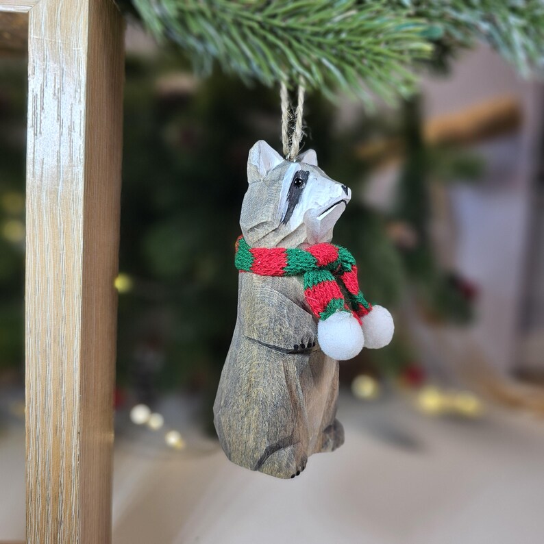 Op de afbeelding: Houten wasbeer ornament met een rood-groene gestreepte sjaal met witte pompoenen. Het ornament is een kerstversiering.