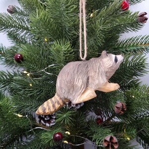 K&ouml;nnte beinhalten: Ein h&ouml;lzerner Waschb&auml;r-Anh&auml;nger mit einem braun-schwarz gestreiften Schwanz, der an einem gr&uuml;nen Weihnachtsbaum h&auml;ngt.
