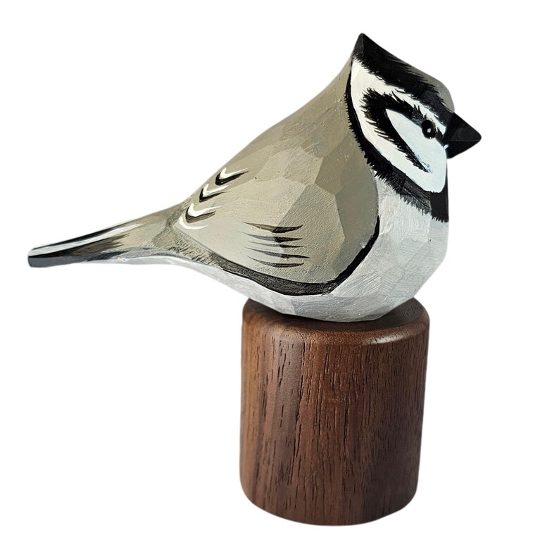 May include: A hand-carved wooden bird figurine with a light gray body, black and white head markings, and a black beak. The bird is perched on a small, round, dark brown wooden base. The figurine has a detailed, faceted design.