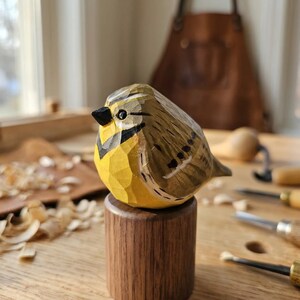 Hand-carved wooden Western Meadowlark figurine with a faceted whittled texture and hand-painted yellow and black plumage. The bird sculpture is perched on a cylindrical wood base in a workshop setting with a leather apron, perfect for rustic home decor, a nature-themed shelf sitter, or a unique gift for bird watchers.