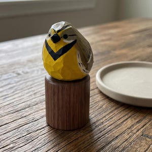 Hand-carved wooden Western Meadowlark figurine with a faceted whittled texture and hand-painted yellow plumage. The bird sculpture is perched on a dark wooden base next to a small white ceramic dish, perfect for rustic home decor, a nature-themed shelf sitter, or a unique gift for bird watchers.