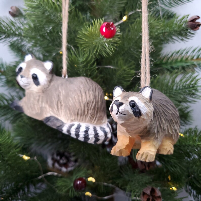 K&ouml;nnte beinhalten: Zwei h&ouml;lzerne Waschb&auml;r-Anh&auml;nger, die an einem Weihnachtsbaum h&auml;ngen. Der Waschb&auml;r links liegt mit seinem Schwanz um seinen K&ouml;rper gewickelt. Der Waschb&auml;r rechts steht mit seinen Pfoten auf dem Boden. Beide Waschb&auml;ren sind in Braun-, Schwarz- und Wei&szlig;t&ouml;nen bemalt.