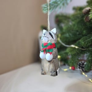 Op de afbeelding: Houten kerstversiering in de vorm van een wasbeer die een rood-groene gestreepte sjaal draagt. De wasbeer houdt twee witte pompoenen in zijn poten.