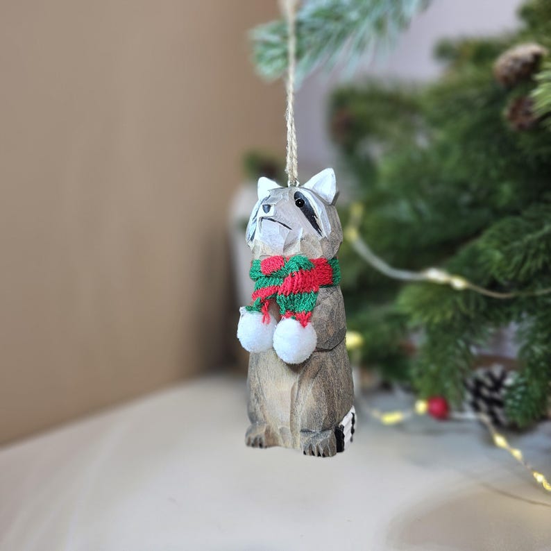Op de afbeelding: Houten kerstversiering in de vorm van een wasbeer die een rood-groene gestreepte sjaal draagt. De wasbeer houdt twee witte pompoenen in zijn poten.
