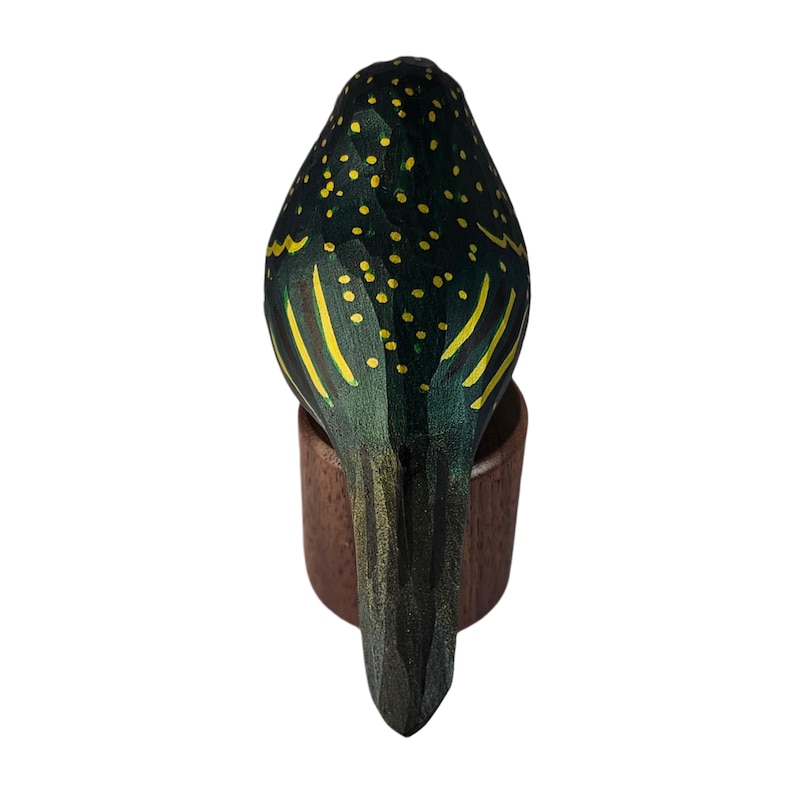 May include: A hand-carved wooden bird figurine, painted in dark green with yellow accents. The bird is perched on a small, round, brown wooden base. The bird's back is detailed with yellow spots and stripes.