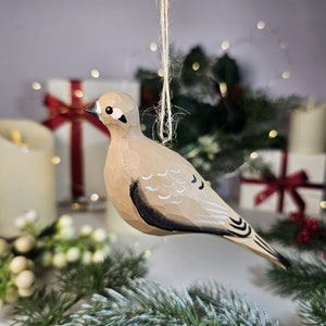 Op de afbeelding: Een handgesneden houten vogelornament, beschilderd in tinten bruin, zwart en wit, hangt aan een touwtje. De vogel heeft een gedetailleerd, getextureerd oppervlak en is ontworpen voor vakantiedecoratie.