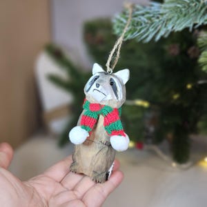 Hand-carved wooden raccoon ornament wearing a red and green knit winter scarf, hanging on a tree. Basswood woodland animal holiday decor.
