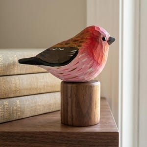 May include: A hand-carved wooden bird sculpture with a red and pink body, brown wings, and a black beak. The bird sits atop a small, cylindrical wooden base. The sculpture is displayed on a dark wooden shelf.