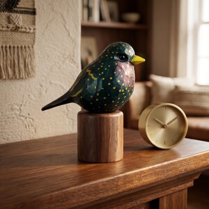 Side profile view of a hand-carved wooden European Starling figurine showcasing its intricate, hand-painted yellow speckles over glossy dark iridescent green and purple plumage. The realistic basswood bird sculpture features a bright yellow beak and is perched on a round black walnut base, displayed on a wooden table next to a circular gold mantel clock. Perfect for nature-inspired home decor, office desk styling, or a unique gift for bird watchers.