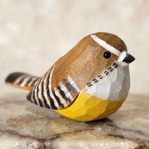 Könnte beinhalten: Eine handgeschnitzte Vogelfigur aus Holz mit braunem Rücken, schwarz-weiß gestreiften Flügeldetails, weißer Brust und leuchtend gelbem Bauch. Der Vogel hat einen kleinen schwarzen Schnabel, ein schwarzes Auge und einen weißen Streifen über dem Auge.