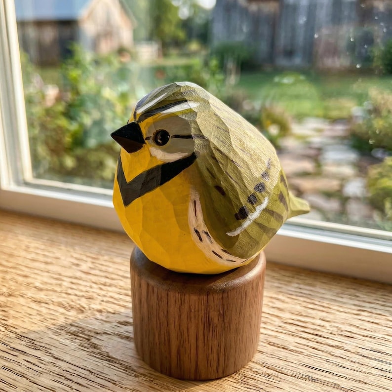 Hand-carved wooden Western Meadowlark figurine featuring a faceted whittled texture and hand-painted yellow breast with a signature black V-neckband. The bird sculpture is perched on a rustic wood base against a soft-focus garden background, perfect for nature-inspired home decor, a windowsill accent, or a unique gift for bird watchers.