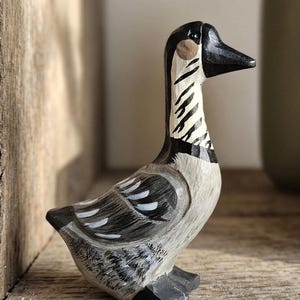May include: A hand-carved wooden goose figurine. The goose has a black head and beak, a cream-colored body with gray and white accents, and black feet. The neck features a distinctive black and white striped pattern. The figurine is displayed on a wooden surface.
